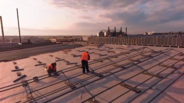 İşçiler gün batımında bir çatıda güneş çiftliği inşa ederler. Erkek yükleyiciler, kırsal kesimdeki bir binanın akşam arkaplanındaki büyük düz çatısına metal temeller atarlar.
