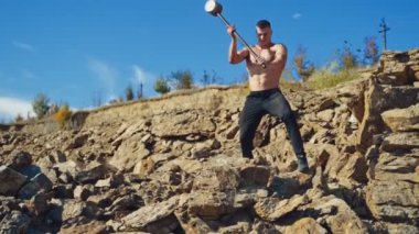Kaslı vücut geliştirici tepede çelik çekiçle antrenman yapıyor. Metal bir aletle dışarıda taş kıran gömleksiz güçlü bir atlet. Yavaş çekim.