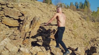 Gömleksiz vücut geliştirici balyozla taş kırıyor. Dağlarda çelik çekiçle taş kıran güçlü bir adam. Yavaş çekim.