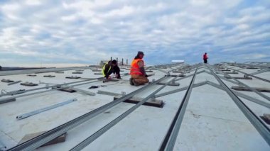 Mavi gökyüzünün altında yenilikçi güneş çiftliğinin kurulması. Çatıdaki güneş panelleri için metal bazlı. Üniformalı işçiler fotovoltaik hücreler için temel oluşturuyorlar.