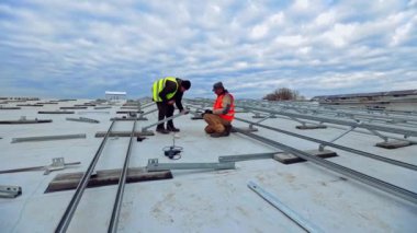 Binanın tepesinde güneş enerjisi çiftliğinin yeni inşaatı. Düz bir çatıya metal taban bağlayan renkli yelekli iki işçi..