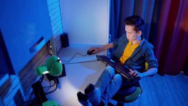 ティーンエイジャーは家庭でコンピュータゲームを楽しんでいる 男の子は机の上に足で椅子に座っている間にコンピュータビデオゲームをしています トップビュー — ストック動画