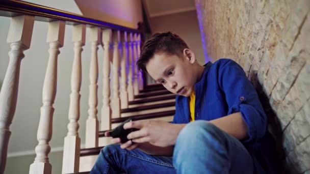 ティーンエイジャーは携帯電話でゲーム中毒です 男の子は階段の上に家に座って 彼の携帯電話でビデオゲームをする — ストック動画