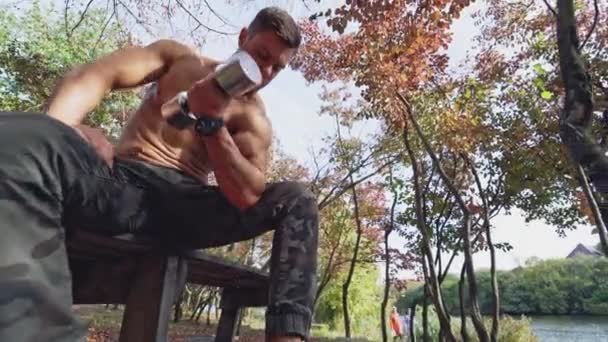 셔츠가없는 운동선수는 덤벨을 야외에서 테이블에 배경에 근육을 아래에서 — 비디오