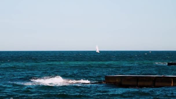 蓝色水底 在海水中的石坞 白色的帆飘浮在遥远的海面上 浪在码头上破浪 形成白色泡沫 慢动作 — 图库视频影像