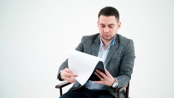 ハンサムなビジネスマンが文書を読んでいる 木製の椅子に座って白い背景に書類を見ている灰色のスーツの若い男 — ストック動画