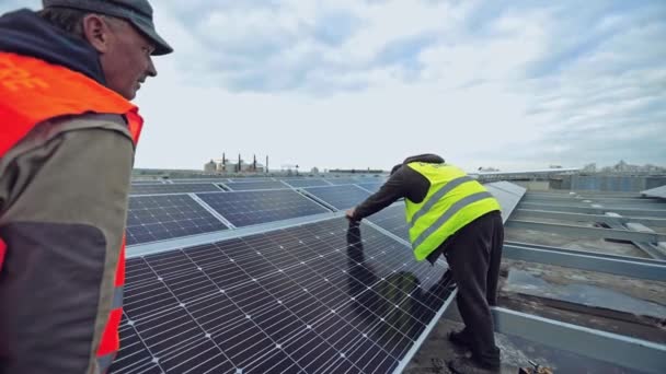 ソーラーパネル設置 現代の太陽光発電所の背景に太陽光発電パネルを取り付ける明るいベストの2人の労働者 オルタナティブエネルギー — ストック動画
