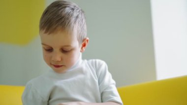 Evde sevimli bir anaokulu çocuğu var. Kanepede oturan ve aşağıya bakan küçük bir çocuğun portresi. Beyaz kazaklı çocuk evde kitap okuyor..