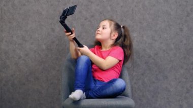 Cep telefonu olan neşeli küçük bir kız. Tişörtlü ve kot pantolonlu şirin bir çocuk stüdyo arka planında izole edilmiş cep telefonuyla selfie çekiyor..