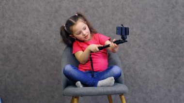 Küçük kız akıllı telefon kullanıyor. Pembe tişörtlü güzel bir çocuk akıllı telefondan selfie çekiyor. Çocuk sandalyede oturuyor ve gri stüdyo arka planında cep telefonuyla poz veriyor..
