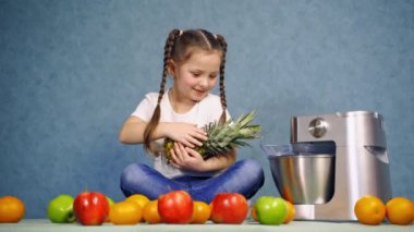 Taze meyveli küçük kız. Masada oturan ve ananasla oynayan sevimli bir çocuk. Organik meyveler ve masada bir sıkma makinesi..
