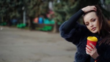 Parktaki şık bir kız. Güneşli bir günde, arka planda plastik bir fincan kahveyle kürklü güzel bir kadının portresi. Yavaş çekim.