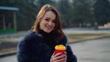 Açık havada kahveyle gülümseyen bir model. Kürk mantolu güzel kız parkta dururken plastik bardaktan kahve içer. Kamerada poz veren genç bir kadın. Yavaş çekim.