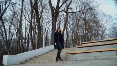 Parktaki güzel kız. Şık bir palto giyen, ağaçların arka planında yapraksız temiz havanın tadını çıkaran esmer kadının portresi. Yavaş çekim.