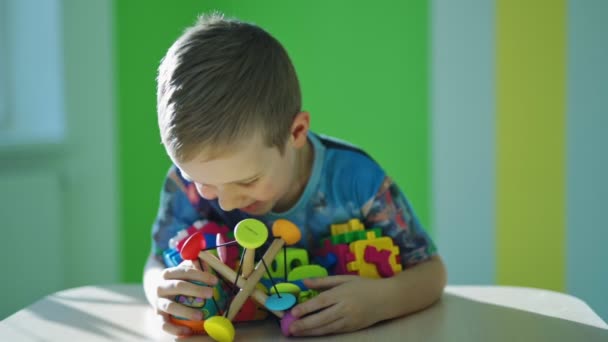 おもちゃを持っている幸せな少年 部屋の背景に多くの明るいおもちゃを持っている青い目を持つ小さな子供の笑顔 ハッピー チャイルド — ストック動画