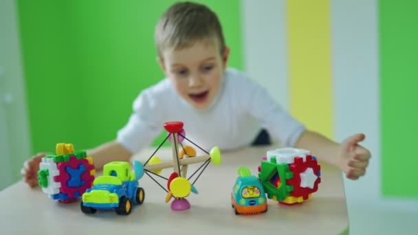 おもちゃを持っている幸せな少年 テーブルの上の明るいおもちゃ かわいい子供は カラフルな部屋の背景にあるすべてのプラスチックのおもちゃをキャプチャします スローモーション — ストック動画