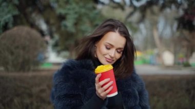 Gülümseyen bir kadının portresi. Kürk mantolu çekici esmer kız parkta plastik bir fincan kahveyle kameraya bakıyor. Yavaş çekim.