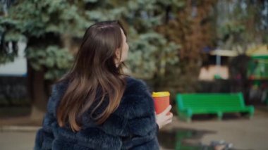 Çekici kız kamerada poz veriyor. Siyah kürklü güzel bir kadın parkta kahve içiyor ve kameraya gülümsüyor..