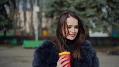 Parktaki çekici genç bayan. Kürklü şık bir kızın yarım boy portresi şehrin arka planında plastik bir fincan kahve tutuyor..