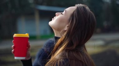Parktaki güzel esmer. Kürk mantolu uzun saçlı kız dışarıda plastik bardak tutuyor. Kamera hareket ediyor.