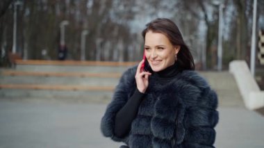 Gülümseyen kadın telefonda konuşuyor. Kürklü neşeli kız parkta duruyor ve cep telefonuyla konuşuyor. Yavaş çekim.