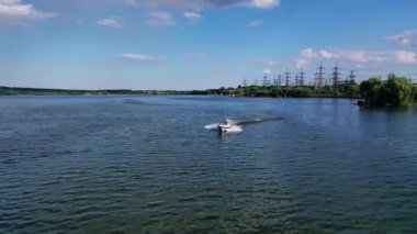 Tekne nehirde yüzüyor. İnsanlar yazın yüksek voltajlı elektrik hatlarının arka planında motorlu tekne ile seyahat ederler. Hava görünümü.