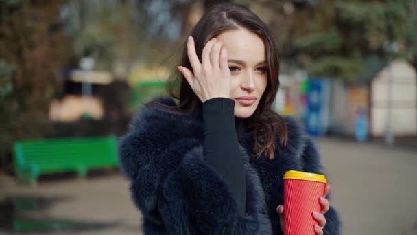 冬のコートで美しい女の子 美しい若い女性は 公園のカメラにコーヒーと笑顔のプラスチックカップを保持しています スローモーション — ストック動画