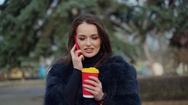 屋外で電話とコーヒーで毛皮のコートを着ている女の子 若い女性の肖像画は 携帯電話を話したり 公園で熱い飲み物を飲んだりする スローモーション — ストック動画