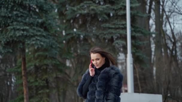公園で携帯を持った高級女の子 公園を歩いている毛皮のコートの魅力的な女性の肖像画と携帯電話の話 スローモーション — ストック動画