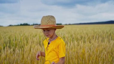 Açık havada gülümseyen bir çocuğun portresi. Hasır şapkalı ve sarı tişörtlü çocuk tarımın içinde dikilip kameraya bakıyor..