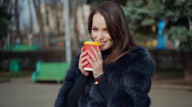 Plastik bardaklı uzun saçlı kız. Kürk mantolu çekici genç kadın şehir parkında kahve içmekten hoşlanıyor. Yavaş çekim.