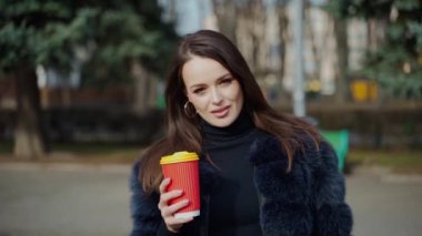 Baştan çıkarıcı kadın portresi. Kabarık ceketli güzel bir kız plastik bardak sıcak içecek tutuyor ve şehir parkında kameraya bakıyor..