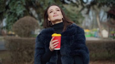 Plastik kahve fincanıyla göz alıcı bir kızın portresi. Parkta dikilip kameraya gülümseyen lüks kürklü çekici genç manken. Yavaş çekim.