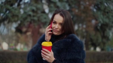 Kürk mantolu çekici bir genç kız. Paltolu güzel bir kadın telefonda konuşuyor ve parkta kahve bardağı tutuyor..