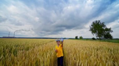 Çocuk dışarıda oynuyor. Tatlı çocuk sarı alanda yürüyor ve yazın bulutlu gökyüzünün altında oyuncak uçağıyla uçuyormuş gibi yapıyor..