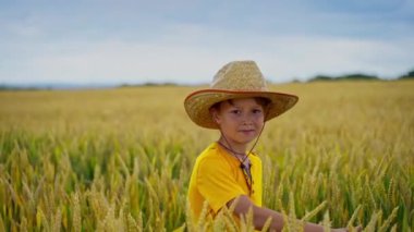 Açık havada küçük bir çiftçi. Hasır şapkalı neşeli çocuk buğday tarlasında duruyor ve kameraya bakıyor. Tarım arazisindeki bir çocuğun portresi.