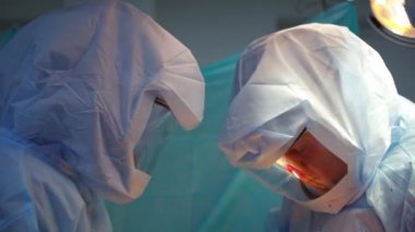 Koruyucu giysili doktorlar. Klinikte beyaz güvenlik üniforması uzmanları hastayı ameliyat ediyor. Maskeli cerrahlar tıbbi aletlerle çalışırlar..