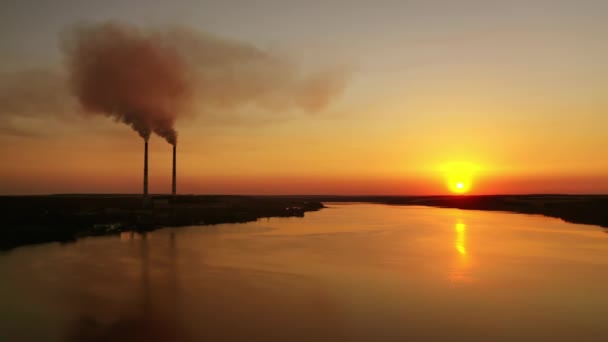 夜の川の近くに工場がある パイプからの汚れた煙は 設定された太陽に対して空中に入ります オレンジ色の空の背景にある湖の近くにある工場のシルエット — ストック動画