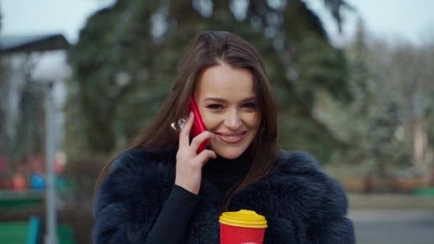 ふわふわのコートでチャーミングな女の子 美しい若いブルネットは携帯電話を話し ぼやけた公園の背景に微笑みます 毛皮のコートの女性は屋外でコーヒーのプラスチック カップを握ります — ストック動画