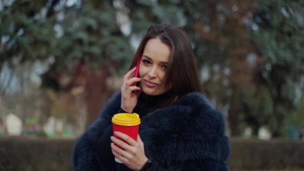迷人的年轻姑娘穿着毛皮大衣 美丽的女人穿着时髦的外套 一边打电话一边拿着咖啡杯在公园里 — 图库视频影像