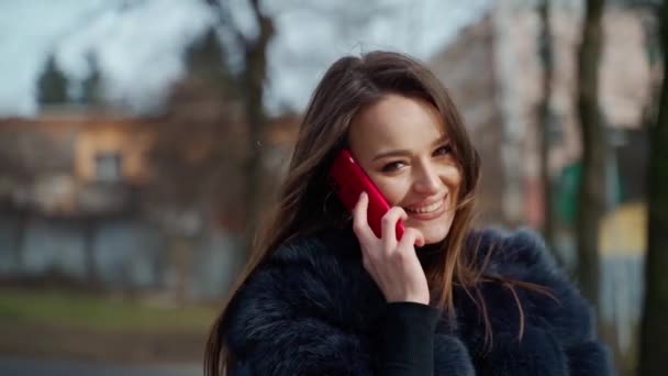 電話を持った笑顔の少女の肖像 毛皮のコートを着た美しい若い女性は魅惑的にカメラのためにポーズし 市の公園で電話を話します 周りのモーションカメラ — ストック動画