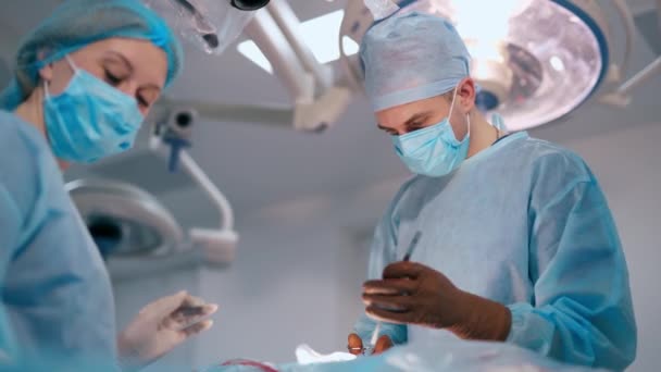 外科医生完成了手术 医生和一名戴蓝色口罩的女护士在明亮的医疗灯下做手术 手术室的专家 — 图库视频影像