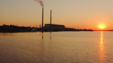 Elektrik santralinin arka planında akşam nehri. Endüstriyel borulardan çıkan duman, batan turuncu güneşe karşı havaya karışır. Gölün yakınındaki bir fabrikanın silueti..