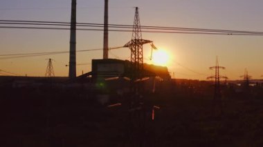 Gün batımında iletim hattının silueti. Fabrika arka planında çelik voltaj elektrik kulesi. Endüstriyel fabrikanın yanındaki uzun elektrik direği. Kamera aşağı iniyor. Kirlilik.