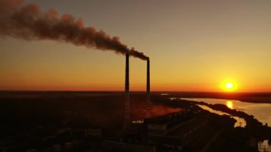 Hava kirliliği. Günbatımında endüstriyel borulardan siyah duman çıkıyor. Nehir kenarındaki zararlı fabrikanın silueti batan güneşe karşı.