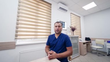 Online danışmanlık. Mavi üniformalı profesyonel erkek doktor masaya oturmuş kameraya bakıyor ve sağlık hizmetleri hakkında konuşuyor..