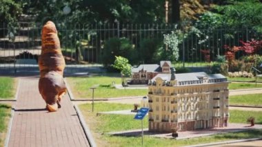 Dışarıdaki komik ejderha kostümlü adam. Güneşli bir günde, oyun dolu binalar boyunca yaz parkında yürüyen dinozorun arka manzarası..