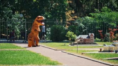Dinozor kostümü parkta yürüyor. Ejderha kostümü giymiş biri, parlak bir yaz gününde el yapımı binaları olan küçük peri şehrinde yürüyor..