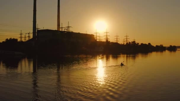 河畔一家工厂的轮廓 日落时 船里的人拿着桨散步 靠近水边的高压电塔挡住落日 — 图库视频影像