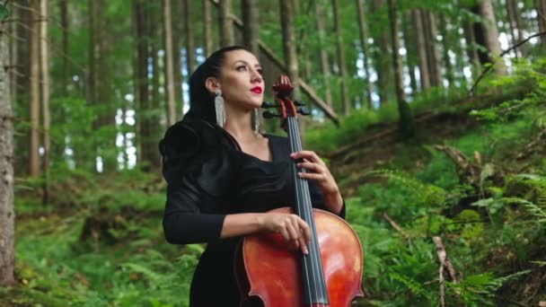 森林里漂亮的大提琴演奏家穿着黑色衣服的奢华女人在绿色大自然的背景下弹奏大提琴 吸引人的女音乐家在户外表演音乐 — 图库视频影像